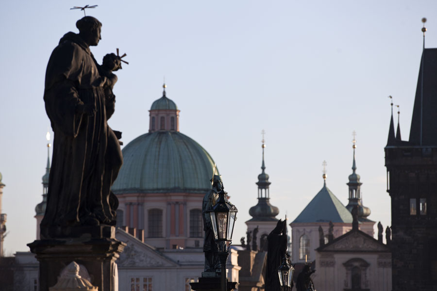 Statue of St. Anthony of Padua and other statues and lanterns on Charles Bridge with the cupola of St. Francis of Assisi church | Ponte Carlo | Cechia