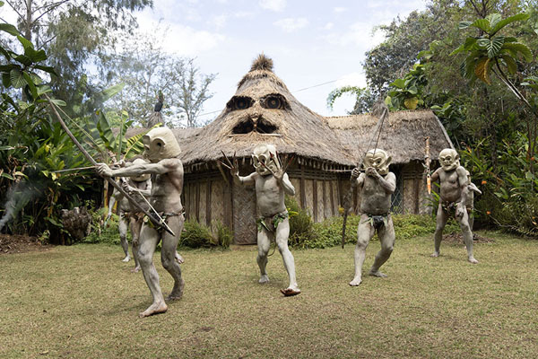 Foto van Asaro mudmen with bows and arrows in front of a typical houseAsaro Mudmen - Papoea Nieuw Guinea