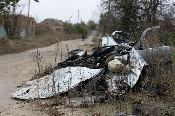 Foto van One of the few inhabitants of Kamyanka on an empty road in Kamyanka with one of the many car wrecks in the foregroundIzyum - Oekraïne